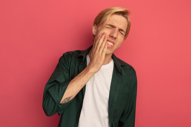 Unpleased with closed eyes young blonde guy wearing green t-shirt putting hand on aching tooth