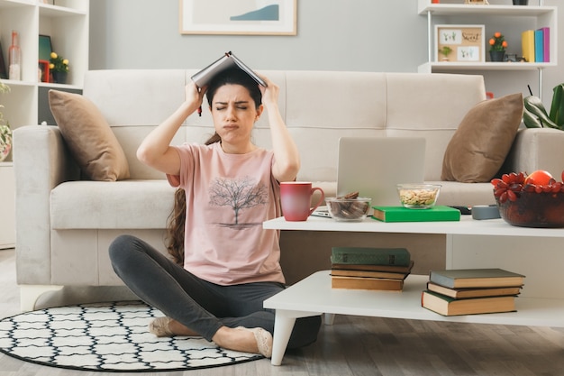 Unpleased with closed eyes young girl covered head with notebook sitting on floor behind coffee table in living room