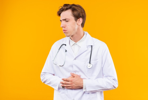 Free photo unpleased with closed eyes young male doctor wearing medical robe with stethoscope grabbed aching stomach isolated on orange wall