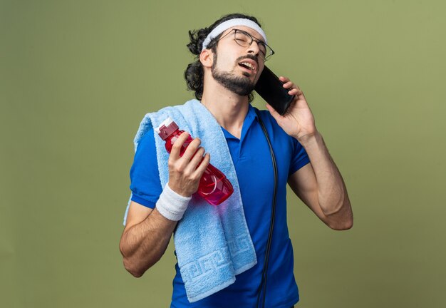 Unpleased with closed eyes young sporty man wearing headband with wristband and towel on shoulder speaks on phone holding water bottle 