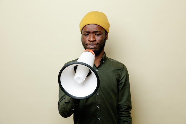 Unpleased young african american male in hat wearing green shirt speaks on loudspeaker isoloated on white background