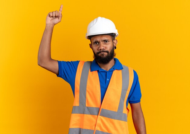 Unpleased young afro-american builder man in uniform with safety helmet pointing up isolated on orange background with copy space