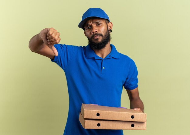 Недовольный молодой афро-американский курьер, держащий коробки для пиццы и листающий вниз, изолирован на оливково-зеленом фоне с копией пространства