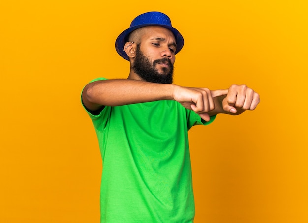Unpleased young afro-american guy wearing party hat showing wrist clock gesture 