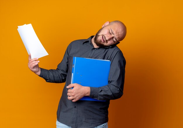 Unpleased young bald call center man holding folder and documents and talking on phone holding phone on shoulder isolated on orange background with copy space