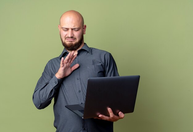 Free photo unpleased young bald call center man holding and looking at laptop and doing no gesture isolated on olive green background with copy space