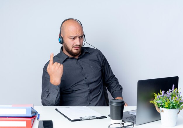 不機嫌そうな若いハゲのコールセンターの男性が机に座って作業ツールで指を上げ、白い背景で隔離のラップトップを見ている
