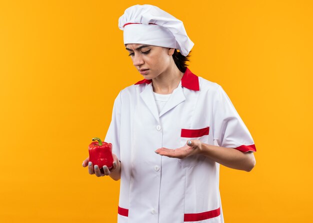 Foto gratuita giovane bella ragazza scontenta in uniforme da chef che tiene e punta con la mano al pepe