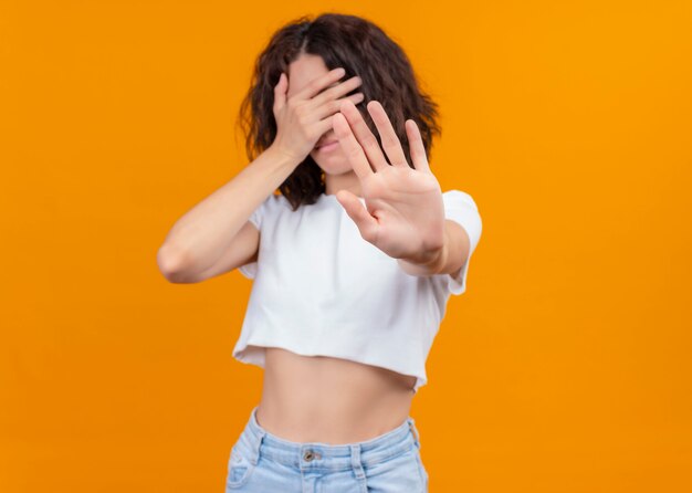 Unpleased young beautiful girl closing her eyes with hand and gesturing stop at camera on isolated orange background with copy space