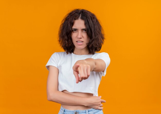 Giovane bella ragazza insoddisfatta che indica alla macchina fotografica e che tiene la sua pancia su fondo arancio isolato con lo spazio della copia