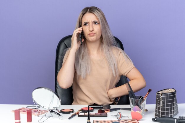 Unpleased young beautiful girl sits at table with makeup tools speaks on phone holding comb putting hand on hip isolated on blue background