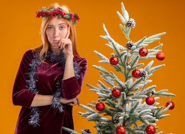 オレンジ色の壁に隔離された頬に手を置き、赤いドレスと花輪を首に付けた花輪を着てクリスマス ツリーの近くに立っている不愉快な美しい少女