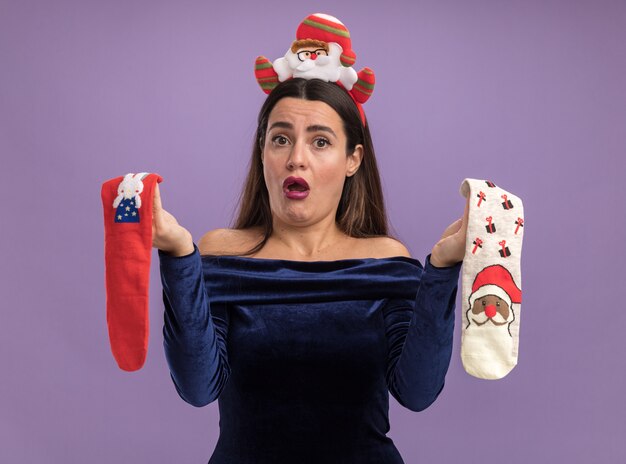 Unpleased young beautiful girl wearing blue dress and christmas hair hoop holding christmas socks isolated on purple background