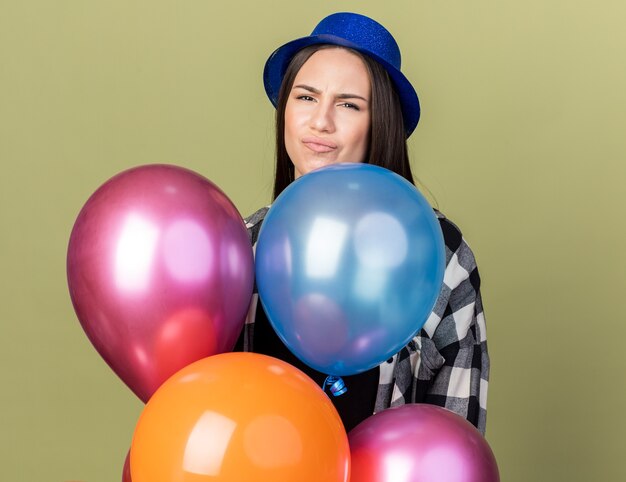 Недовольная молодая красивая девушка в синей шляпе стоит за воздушными шарами, изолированными на оливково-зеленой стене