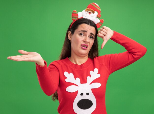 緑の壁に分離された側の手でポイントを親指を下に示すクリスマスの髪のフープとクリスマス セーターを着ている不愉快な美しい少女