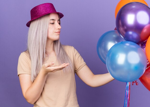 Недовольная молодая красивая девушка в партийной шляпе держит и указывает рукой на воздушные шары