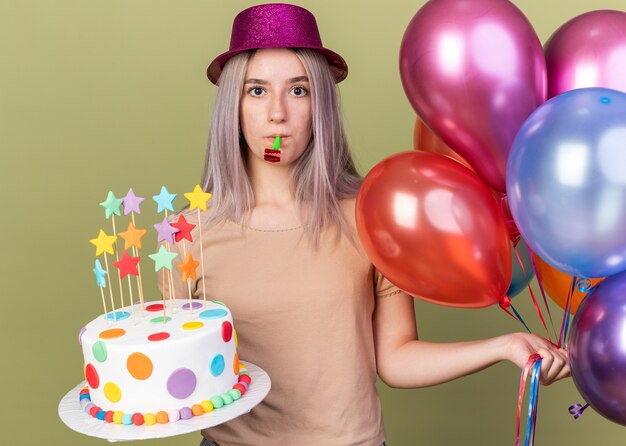 オリーブグリーンの壁に分離されたケーキを吹くパーティー笛と風船を保持しているパーティー帽子をかぶって不機嫌そうな若い美しい少女