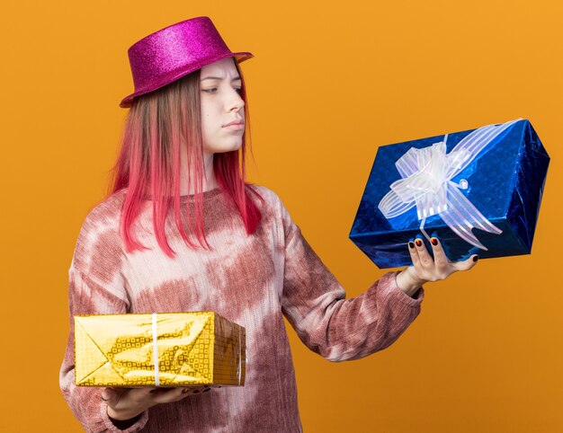 Unpleased young beautiful girl wearing party hat holding and looking at gift boxes isolated on orange wall