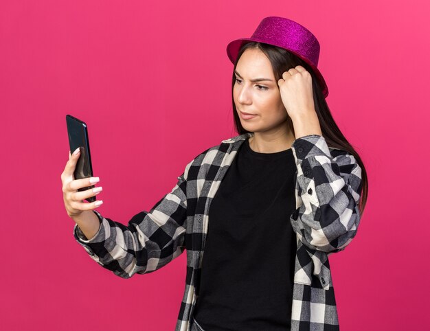 Unpleased young beautiful girl wearing party hat holding and looking at phone 