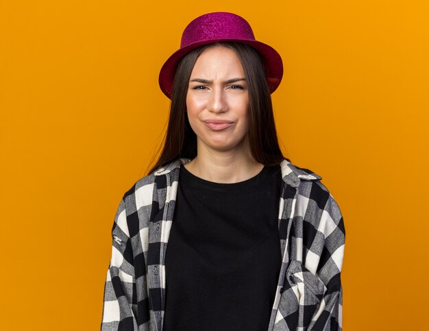 Unpleased young beautiful girl wearing party hat isolated on orange wall