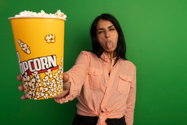 Бесплатное фото Недовольная молодая красивая девушка в розовой футболке держит ведро попкорна, показывая язык, изолированный на зеленом