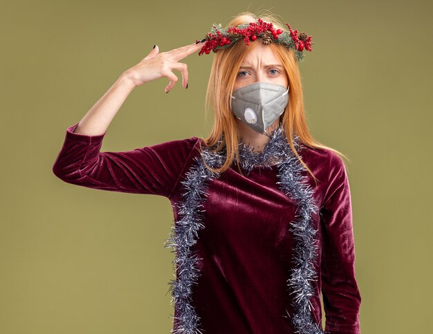 Unpleased young beautiful girl wearing red dress with wreath and medical mask with garland on neck showing suicide with pistol gesture isolated on olive green background