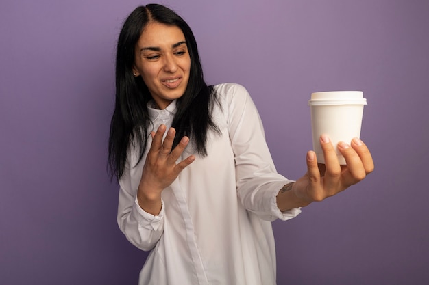 Недовольная молодая красивая женщина в белой футболке держит и указывает рукой на чашку кофе