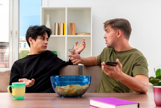 unpleased young blonde and brunette handsome guys sit at table with raised hands looking at each other blonde man holding phone inside living room