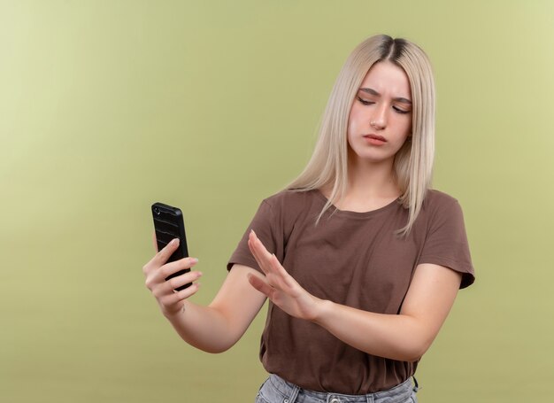 Unpleased young blonde girl holding mobile phone looking at it and gesturing no on isolated green wall with copy space