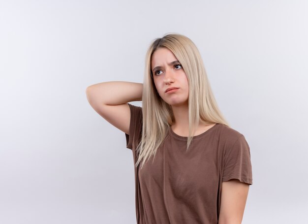 Unpleased young blonde girl putting hand behind her head looking at left side on isolated white wall with copy space