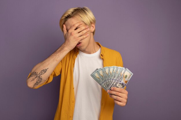 Unpleased young blonde guy wearing yellow t-shirt covered face with hand and holding cash isolated on purple