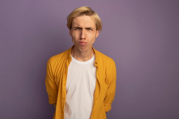 Free photo unpleased young blonde guy wearing yellow t-shirt putting hands on waist isolated on purple with copy space