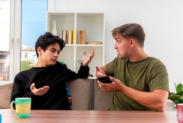 Free photo unpleased young blonde handsome man holds and points at phone with hand sitting at table and looking at clueless young brunette handsome boy