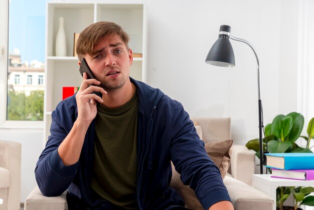 Unpleased young blonde handsome man sits on armchair talking on phone