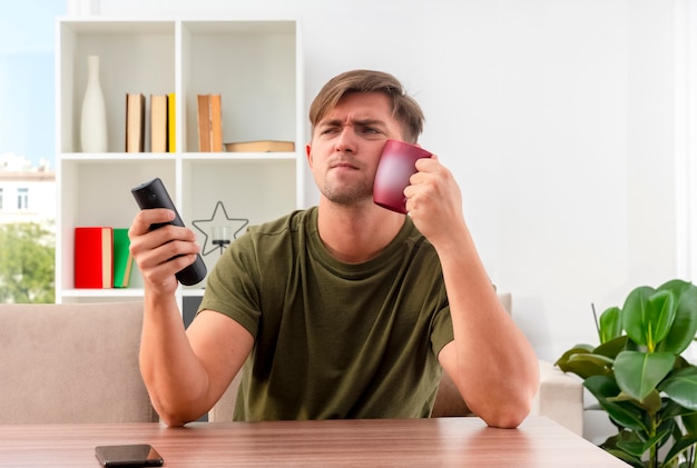 Unpleased young blonde handsome man sits at table holding tv remote and putting cup in face