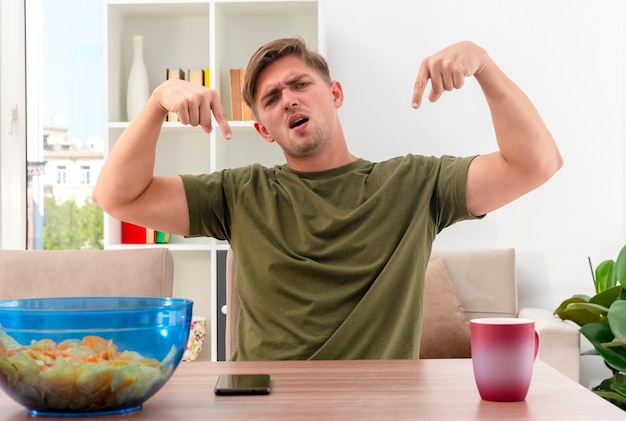 Unpleased young blonde handsome man sits at table with bowl of chips phone and cup pointing down with two hands inside the living room