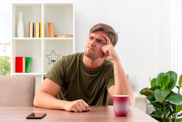 Unpleased young blonde handsome man sits at table with cup and phone