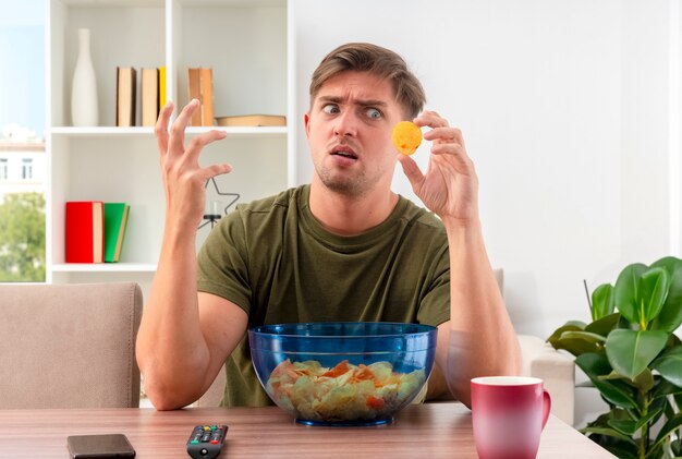 불쾌한 젊은 금발의 잘 생긴 남자는 칩의 전화 그릇과 손을 들고 거실 내부의 칩을보고있는 테이블에 앉아있다.