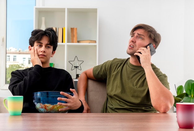 unpleased young blonde handsome man talking on phone sitting at table with disappointed young brunette handsome guy putting hand on face holding bowl of chips inside design living room