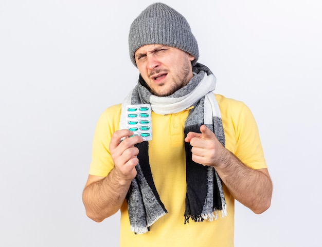 Unpleased young blonde ill man wearing winter hat and scarf holds pack of medical pills and points at front isolated on white wall