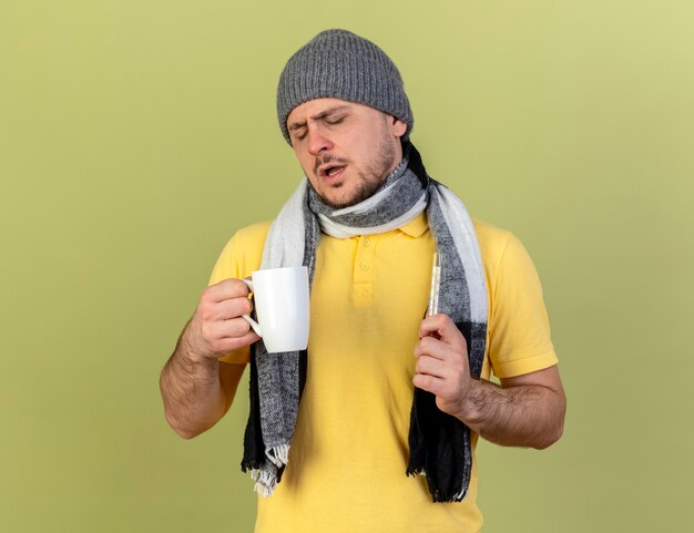 Unpleased young blonde ill slavic man wearing winter hat and scarf holds cup and thermometer isolated on olive green wall with copy space
