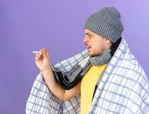 格子縞のホールドに包まれた冬の帽子とスカーフを身に着けている不機嫌な若い金髪の病気のスラブ人は、コピースペースで紫色の壁に隔離された温度計を見て