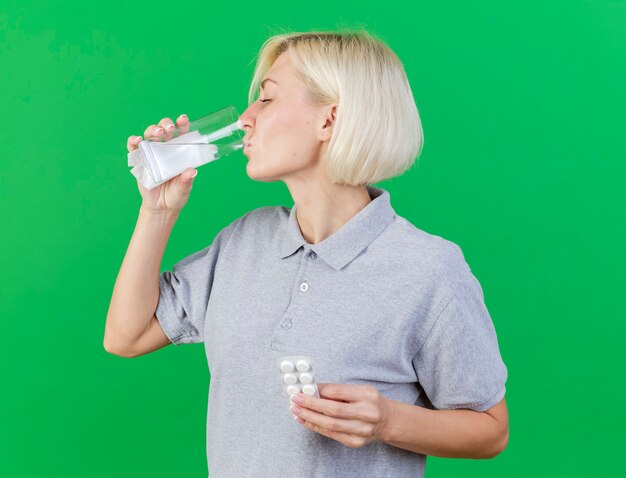 Unpleased young blonde ill slavic woman drinks glass of water and holds pack of medical pills isolated on green wall with copy space
