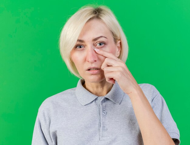 Unpleased young blonde ill slavic woman puts finger on eyelid isolated on green