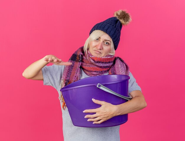 Недовольная молодая блондинка больная славянская женщина в зимней шапке и шарфе держит руку над пластиковым ведром, изолированным на розовой стене с копией пространства