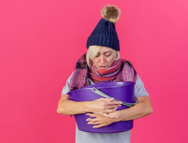 Unpleased young blonde ill slavic woman wearing winter hat and scarf holds and looks at plastic bucket having nausea isolated on pink wall with copy space