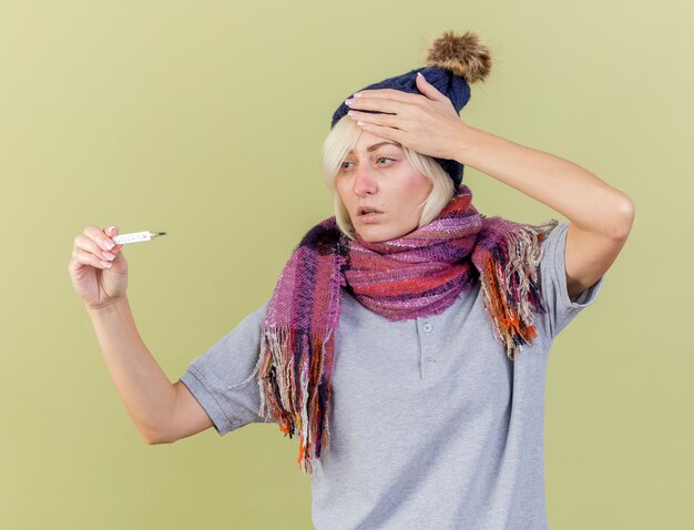 Unpleased young blonde ill slavic woman wearing winter hat and scarf puts hand on head looking at thermometer isolated on olive green wall with copy space