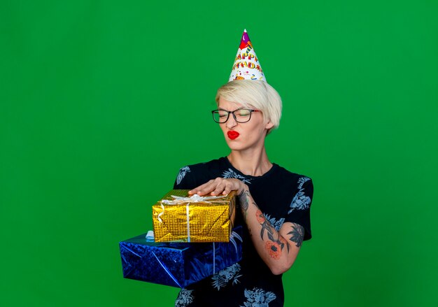 Недовольная молодая блондинка тусовщица в очках и кепке на день рождения держит подарочные коробки с закрытыми глазами, изолированными на зеленом фоне с копией пространства