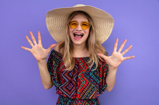 Unpleased young blonde slavic girl with sunglasses and with sun hat standing with raised hands