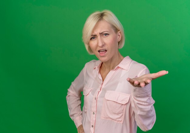 Unpleased young blonde slavic woman showing empty hand and putting another one on waist isolated on green background with copy space